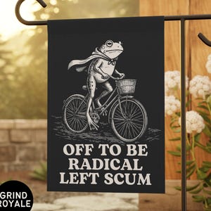May include: A black garden flag featuring a white line art illustration of a frog wearing a cape and riding a bicycle. The text below the frog reads OFF TO BE RADICAL LEFT SCUM. The flag is displayed on a black metal stand in an outdoor setting.