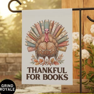 May include: A rectangular garden flag featuring a central turkey with a tail made of colorful books. Below the turkey are autumn leaves and acorns. The text THANKFUL FOR BOOKS is printed in brown at the bottom on a light background.