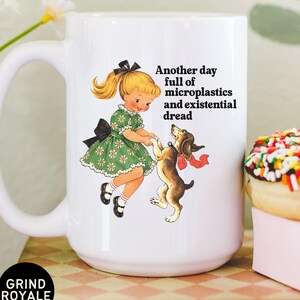 May include: A white ceramic mug featuring a vintage-style illustration of a blonde child in a green dress dancing with a brown dog. The text reads: Another day full of microplastics and existential dread. A black circular logo says GRIND ROYALE.