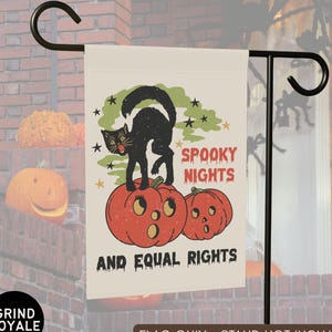 Può includere: Una bandiera da giardino rettangolare con sfondo color crema raffigurante un gatto nero arcuato sopra due zucche di Halloween arancioni. Il testo recita SPOOKY NIGHTS AND EQUAL RIGHTS in lettere rosse e nere, accentuato da fumo verde e stelle nere.