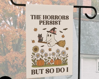 May include: A rectangular garden flag with a cream background featuring the text THE HORRORS PERSIST BUT SO DO I. The illustration shows a ghost wearing a hat riding a broom with a black cat, surrounded by sunflowers, daisies, pumpkins, and bats.