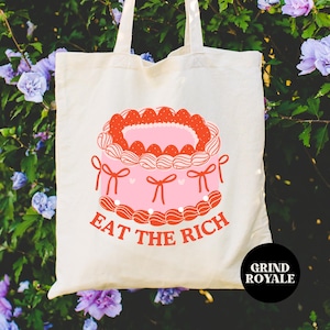May include: A white canvas tote bag with a pink and red cake design and the text "Eat the Rich". The bag has a black tag with the text "Grind Royale".
