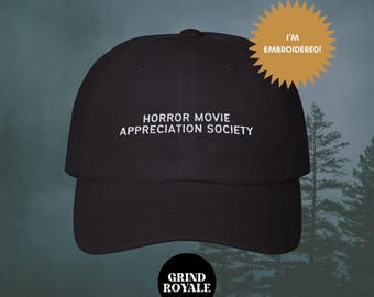 Könnte beinhalten: Schwarze Baseballkappe mit weiß gesticktem Text, der "Horror Movie Appreciation Society" lautet. Die Kappe hat einen gebogenen Schirm und ist vor einem nebligen Waldhintergrund zu sehen.