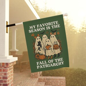 May include: Green Halloween flag with the text "MY FAVORITE SEASON IS THE FALL OF THE PATRIARCHY." The flag features illustrations of ghosts, pumpkins, bats, and fall foliage. A black cat is held by one of the ghosts.