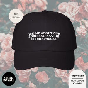 May include: Black baseball cap with the text "ASK ME ABOUT OUR LORD AND SAVIOR PEDRO PASCAL" in white. The cap has a curved brim and a rounded crown. The words "THE ORIGINAL", "GRIND ROYALE", "EMBROIDERED", and "MORE COLORS AVAILABLE" are also visible.