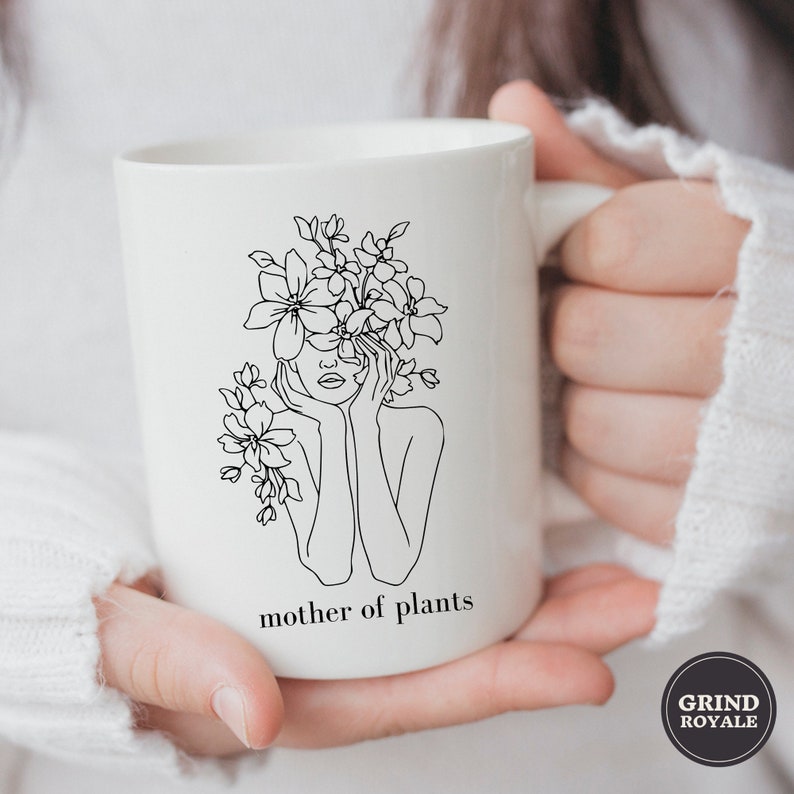 May include: White ceramic mug with a black line drawing of a woman with flowers covering her face. The text "mother of plants" is printed below the drawing.