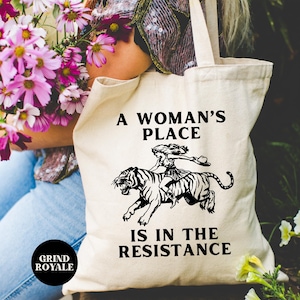 May include: A white canvas tote bag with black text that reads "A woman's place is in the resistance". A black and white illustration of a woman riding a tiger is above the text.