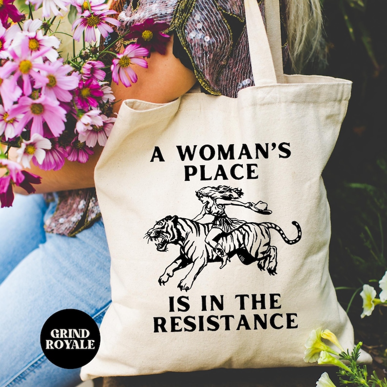 May include: A white canvas tote bag with black text that reads "A woman's place is in the resistance". A black and white illustration of a woman riding a tiger is above the text.
