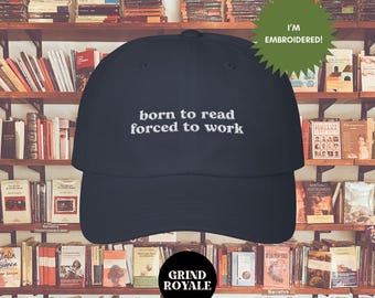 Pode incluir: Um boné de beisebol azul marinho com texto branco bordado que diz "nascido para ler, forçado a trabalhar". O boné está na frente de uma estante cheia de livros.