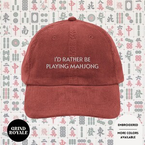 Peut inclure: Casquette de baseball en velours côtelé de couleur rouille avec le texte brodé "I'D RATHER BE PLAYING MAHJONG". La casquette a une visière incurvée et une couronne arrondie. Le fond présente un motif de tuiles de Mahjong.
