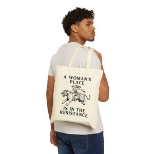 May include: A white canvas tote bag with black text and a graphic of a woman riding a tiger. The text reads "A Woman's Place Is In The Resistance."