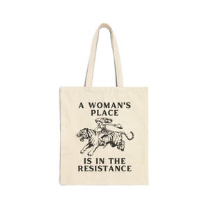 May include: A beige tote bag with a black graphic of a woman riding a tiger. The text "A Woman's Place Is In The Resistance" is printed on the bag.