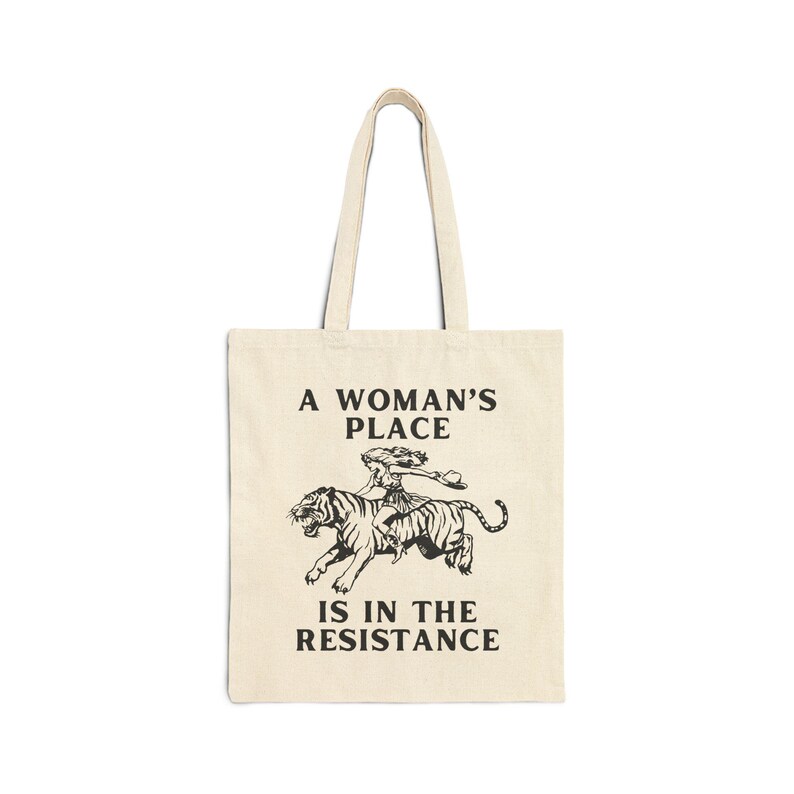 May include: A beige tote bag with a black graphic of a woman riding a tiger. The text "A Woman's Place Is In The Resistance" is printed on the bag.