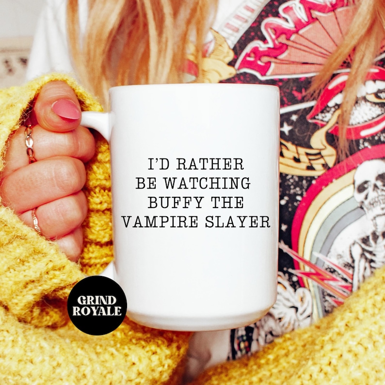 May include: White ceramic mug with black text that reads "I'd rather be watching Buffy the Vampire Slayer". The mug is held by a person wearing a yellow sweater.