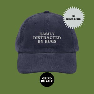 Puede incluir: Una gorra de béisbol de pana azul marino con texto blanco bordado que dice "EASILY DISTRACTED BY BUGS". La gorra tiene un logotipo en blanco y negro que dice "GRIND ROYALE" en la parte inferior.