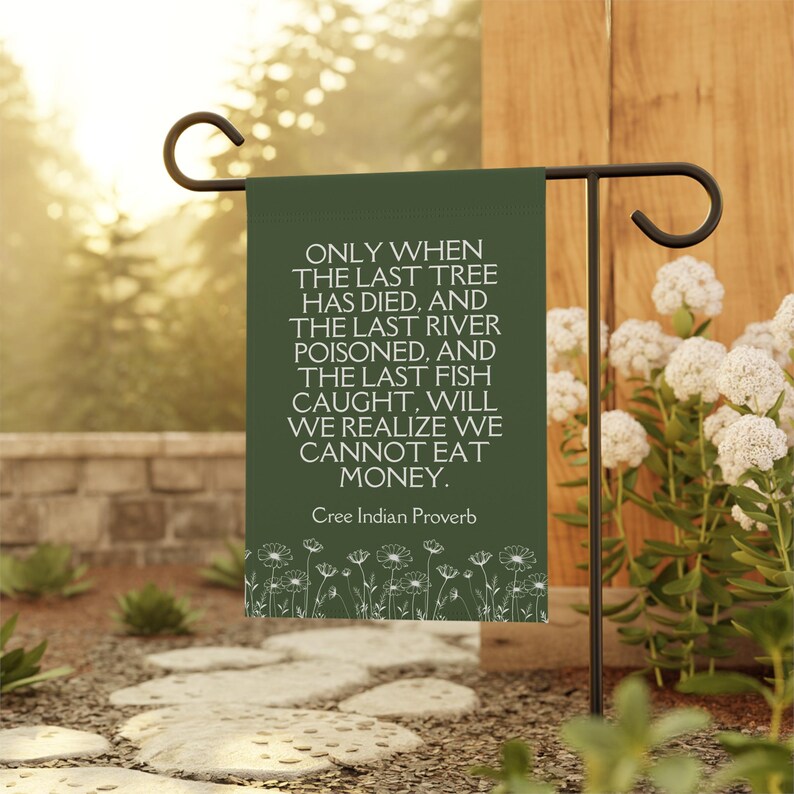 May include: A green garden flag with white text and a white floral border. The text reads: 'ONLY WHEN THE LAST TREE HAS DIED, AND THE LAST RIVER POISONED, AND THE LAST FISH CAUGHT, WILL WE REALIZE WE CANNOT EAT MONEY. Cree Indian Proverb'