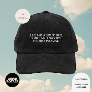 Peut inclure: Casquette de baseball en velours côtelé noir avec le texte blanc brodé : "ASK ME ABOUT OUR LORD AND SAVIOR PEDRO PASCAL". La casquette comporte également les textes "THE ORIGINAL", "GRIND ROYALE", "EMBROIDERED", et "MORE COLORS AVAILABLE".