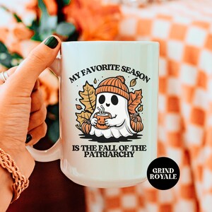 May include: White ceramic mug with a ghost wearing a hat and holding a cup of coffee. The mug reads "My favorite season is the fall of the patriarchy" and "Grind Royale" at the bottom.