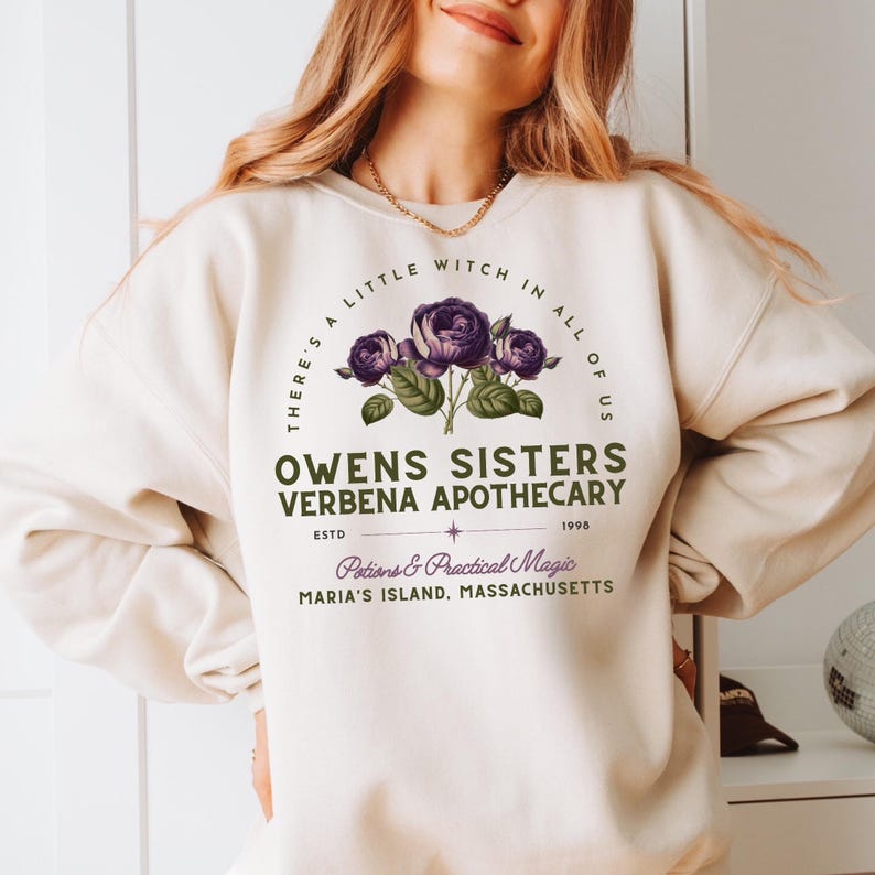 May include: Cream-colored sweatshirt featuring a floral design with purple roses and green leaves. The text reads "THERE'S A LITTLE WITCH IN ALL OF US" and "OWENS SISTERS VERBENA APOTHECARY" with additional text below. The sweatshirt has a crew neck.