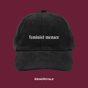 Könnte beinhalten: Eine schwarze Baseballkappe aus Cord mit dem in Weiß gestickten Text "feminist menace" in einer gotischen Schriftart auf der Vorderseite. Die Kappe hat eine strukturierte Krone, einen gebogenen Schirm und eine sichtbare vertikale Cordstruktur.
