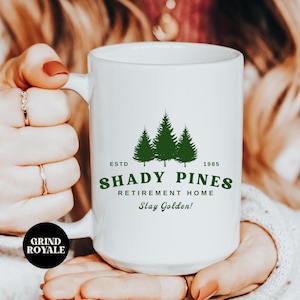 Puede incluir: Taza de cerámica blanca con un logotipo verde que dice "Shady Pines Retirement Home" con el texto "Estd 1985" y "Stay Golden!"