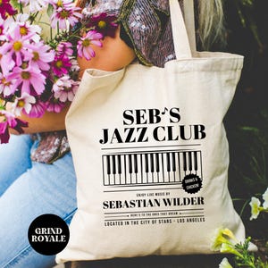 May include: A white canvas tote bag with black text that reads "Seb's Jazz Club" with a black and white piano keyboard graphic. The text "Enjoy live music by Sebastian Wilder" is below the keyboard graphic. A small circle with the text "Drinks & Chicken" is above the keyboard graphic. The text "Here's to the ones that dream" is below the name. The text "Located in the city of stars - Los Angeles" is at the bottom of the bag.