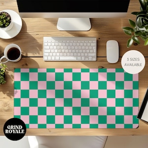 May include: A rectangular desk mat featuring a pink and green chequered design. The mat has the text "3 SIZES AVAILABLE" and "GRIND ROYALE". A keyboard, mouse, and monitor are visible on the wooden desk.
