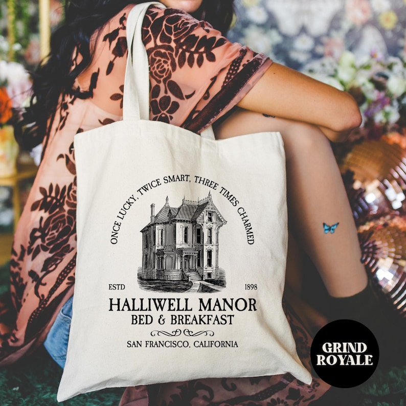 May include: A white canvas tote bag with black text and a black and white illustration of a house. The text reads 'Once lucky, twice smart, three times charmed', 'ESTD 1898', 'Halliwell Manor Bed & Breakfast', and 'San Francisco, California'. The text 'Grind Royale' is also visible in the bottom right corner of the image.