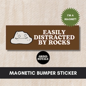 May include: A brown rectangular magnetic bumper sticker with white text that reads "Easily Distracted By Rocks". A cartoon rock with a smiling face is on the left side of the sticker. The text "I'm a Magnet!" is on a green starburst in the upper right corner. The text "Grind Royale" is on a black circle in the lower center of the sticker.