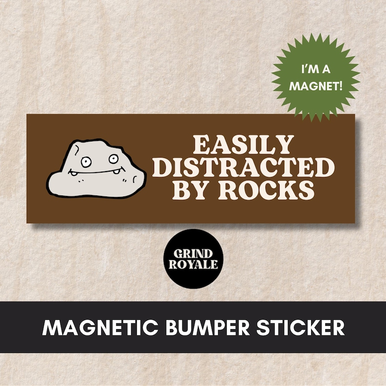 May include: A brown rectangular magnetic bumper sticker with white text that reads "Easily Distracted By Rocks". A cartoon rock with a smiling face is on the left side of the sticker. The text "I'm a Magnet!" is on a green starburst in the upper right corner. The text "Grind Royale" is on a black circle in the lower center of the sticker.