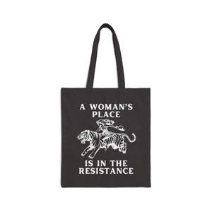 May include: A black tote bag with a white graphic of a woman riding a tiger. The text "A Woman's Place Is In The Resistance" is printed on the bag.