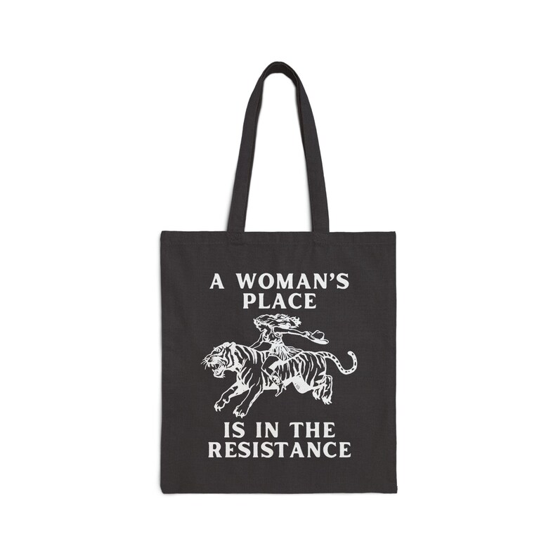 May include: A black tote bag with a white graphic of a woman riding a tiger. The text "A Woman's Place Is In The Resistance" is printed on the bag.