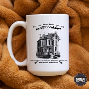 May include: White ceramic mug with a black and white illustration of a house and the text "Owens Sisters Bed & Breakfast Chocolate Cake for Breakfast & Midnight Margaritas Maria's Island, Massachusetts".