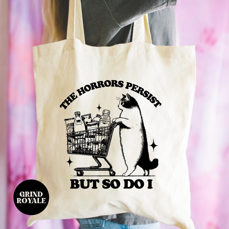 May include: A cream-colored tote bag features a black illustration of a cat pushing a shopping cart filled with groceries.  The text on the bag reads, 'The horrors persist but so do I.' The bag is made of canvas and has a simple design.  The overall theme is dark humor and relatable shopping experiences.