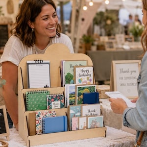 May include: A tiered wooden display stand holding various paper goods, including spiral-bound notebooks, greeting cards with 'Happy Birthday' and 'Thank You' text, and a 'To do list' notepad. The light-coloured wood rack features three shelves.