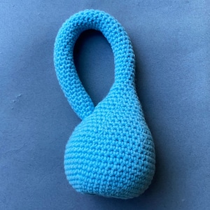 Puede incluir: Un juguete de color azul claro en forma de lágrima tejido a crochet con un lazo en la parte superior.