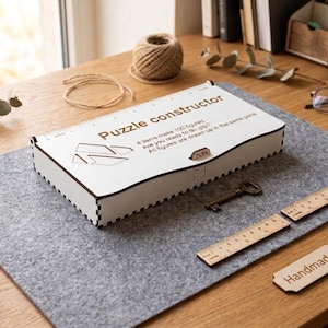以下が含まれることがあります： Puzzle constructorと書かれた白い長方形の木製ボックス。4 items make 100 figured. Are you ready to do gift? All figures are drawn up in the same plane.というテキストが印字されています。ボックスにはプッシュ式の留め具があり、縁はダークブラウンです。
