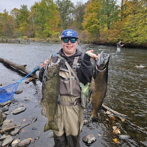 May include: A person standing in a river holding two large fish. The fish are dark in color with visible scales. The person is wearing waders, a black hoodie, and a blue cap. A blue fishing net is visible in the background, along with other people fishing.