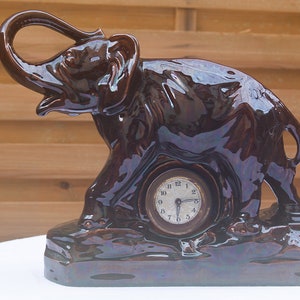 May include: A dark brown ceramic elephant figurine with a clock face embedded in its side. The elephant's trunk is raised and curled, and its eyes are closed. The clock face has black Roman numeral markings and black hands.