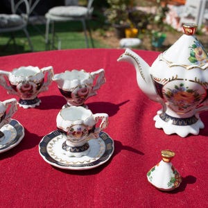 Op de afbeelding: Een miniatuur porseleinen theeservies met bloemmotieven. De set bevat een theepot, een roomkan, een suikerpot en vier theekopjes en schoteltjes. De theeset is geschilderd in tinten blauw, wit en goud.