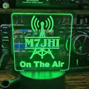 May include: A square, green-lit acrylic sign featuring a radio tower icon, the call sign M7JHI, and the text On The Air. The sign is mounted on a circular black base and is positioned in front of a Yaesu radio transceiver and a Mydel power supply.