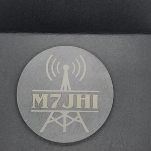 May include: A circular silver sticker featuring a radio tower design and the text "M7 JHI". The sticker is set against a dark grey background, with white and black accents.