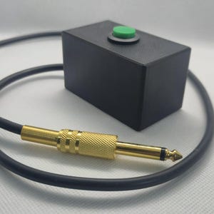 May include: A black rectangular box with a single green circular button on the top surface, connected to a black cable that terminates in a gold-coloured metal quarter-inch audio jack plug with a textured grip section.