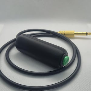 May include: A black cylindrical push-button switch attached to a black cable with a gold-coloured metal jack connector. The switch features a small, round, bright green button on one end, and the cable is coiled in a circular shape.