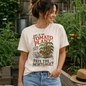 May include: A cream-colored short-sleeve t-shirt featuring a graphic of a tomato plant in a garden bed. The text reads: Why is my tomato plant acting like it pays the mortgage? The design includes red tomatoes, green leaves, and a watering can.