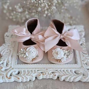 以下が含まれることがあります： 白い花とラインストーンが付いたピンクのベビーシューズのペア。靴にはサテンのリボンが前面にあり、白いフレームの上に置かれています。