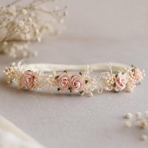 Pode incluir: Uma tiara floral com rosas de tecido em rosa suave, pequenos detalhes de contas brancas e pormenores delicados de renda numa banda fina e flexível. O arranjo é centrado com grupos de rosas e intercalado com pequenos elementos florais.