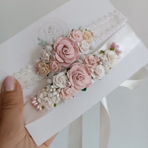 May include: Two wedding garters featuring white lace and satin bands adorned with clusters of artificial dusty pink and cream roses, small white flower buds, and tiny pearl-like beads, presented on a white rectangular display card.