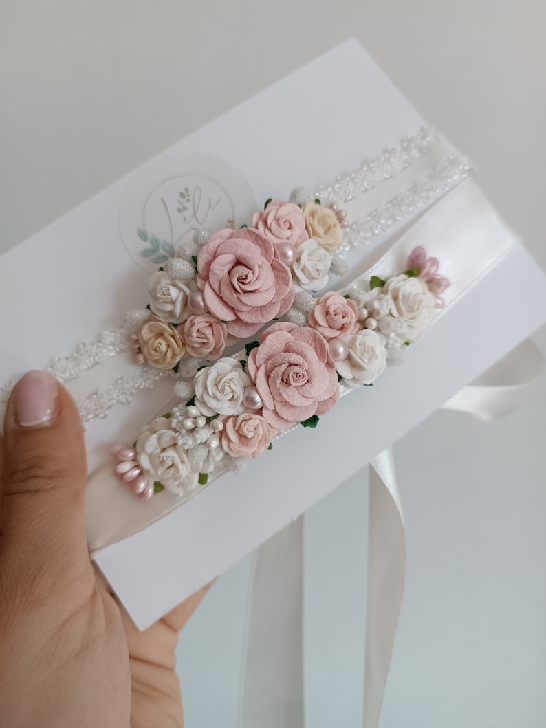May include: Two wedding garters featuring white lace and satin bands adorned with clusters of artificial dusty pink and cream roses, small white flower buds, and tiny pearl-like beads, presented on a white rectangular display card.