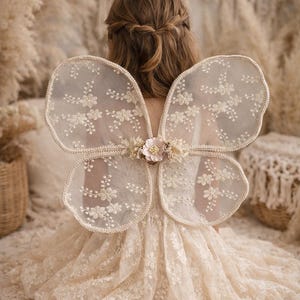 Könnte beinhalten: Ein Kind trägt ein cremefarbenes Spitzenkleid und passende Feenflügel. Die Flügel sind mit floralen Stickereien und einem zarten Besatz verziert. Ein floraler Akzent befindet sich an der Basis der Flügel. Der Hintergrund ist sanft und neutral.